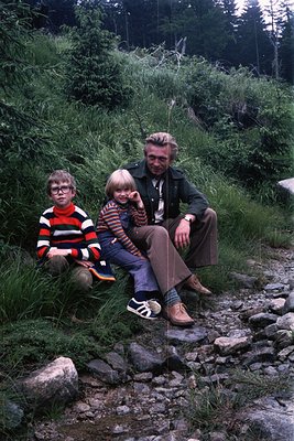 A family of three poses on a rocky forest path, likely 1970s. The man wears a dark jacket, patterned shirt, and wide-brimmed ...