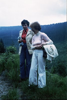 Two women in 1970s outdoor attire pose on a grassy hillside, surrounded by dense forest. The woman on left wears a blazer, bl...