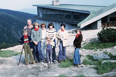 Family group photo in front of a mid-century alpine resort, likely 1970s. Seven individuals pose on a rocky slope near a larg...