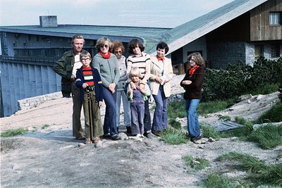 Group portrait of seven individuals (adults + children) in 1970s outdoor setting, likely Bulgaria. Concrete block buildings a...