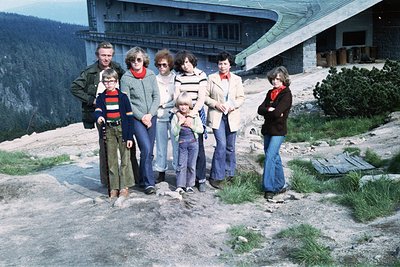 Family group photo in alpine setting, 1970s. Seven individuals pose on rocky terrain near a Brutalist-style building with con...
