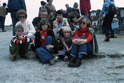 Family group shot on gravel surface, likely a public event or gathering. Adults and children in 1970s attire—wide-legged trou...