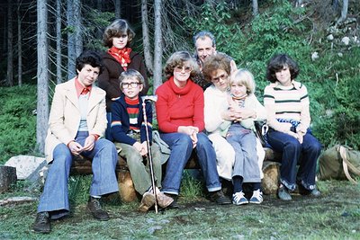 Family group photo in a forested setting, likely 1970s. Eight individuals pose on a wooden bench surrounded by pine trees. Cl...