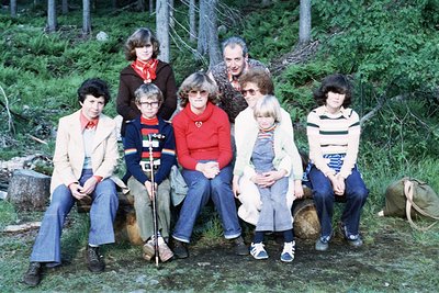 Group portrait of eight individuals in a forested area, likely 1970s. Casual attire includes jackets, scarves, and jeans, sug...