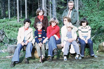 Vintage group photo in a forested area, likely late 1970s–early 1980s. Eight individuals pose on a fallen log, dressed in cas...