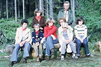 Family group shot in a forested area, 1970s. Eight individuals pose on a wooden bench surrounded by pine trees, wearing 70s f...