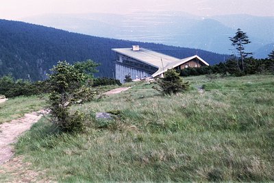 Mid-20th century alpine lodge perched on a grassy plateau, surrounded by dense forest and rugged peaks. Architectural style s...