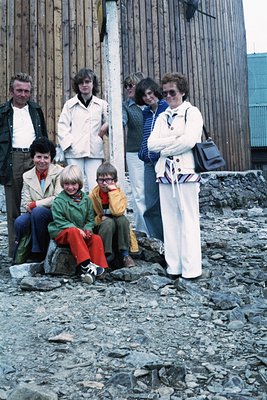 Family portrait in rugged coastal setting, likely 1970s. Seven individuals pose on rocky shore near weathered wooden structur...