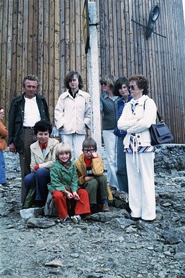 Family portrait in front of a wooden industrial structure, likely a 1970s outdoor setting. Eight individuals pose—adults in b...