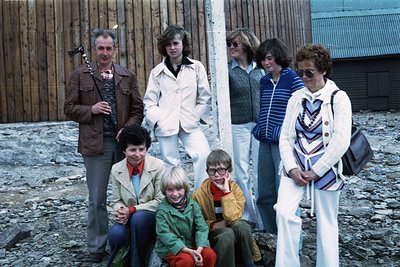 Family portrait featuring seven individuals in 1970s outdoor setting. Wooden fence and rustic building in background. Men in ...