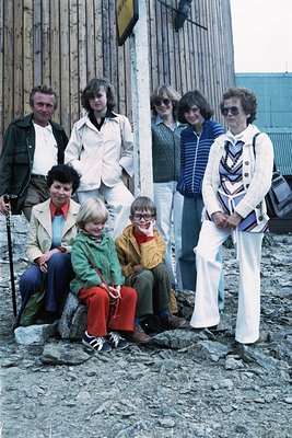 Family portrait from the 1970s, likely Western Europe or North America. Seven individuals pose outdoors on rough gravel near ...