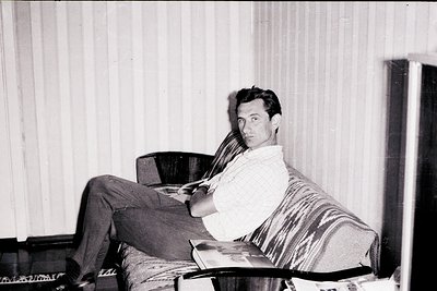 Mid-century man lounges in a striped robe on a modernist armchair, holding a book. Indoor setting with vertical wooden blinds...