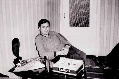 Black-and-white portrait of a seated man in a casual, buttoned-up shirt, leaning on a table with vintage audio equipment—like...