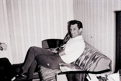 Black-and-white portrait of a man in a relaxed indoor setting, leaning back on a striped armchair. He wears a striped sweater...