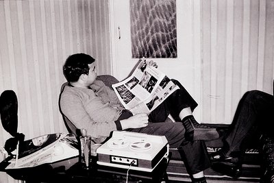 Black-and-white interior shot of a man in a relaxed posture, reading a newspaper while seated in a reclined office chair. Rec...
