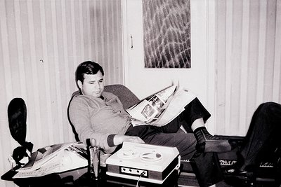 Vintage black-and-white photo of a man engrossed in a large newspaper, seated on a couch in a modest indoor setting. Retro st...