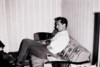 Mid-century man lounges in a striped armchair, holding a pipe and open book. Plain curtained window and minimalist furniture ...