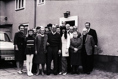 Group portrait in front of a residential building, likely mid-20th century (1950s–1970s). Twelve individuals pose outdoors, d...