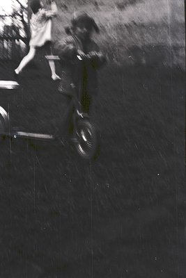 Blurred monochrome street scene capturing motion blur of cyclists on wet pavement, likely post-rain. Reflections on glossy su...