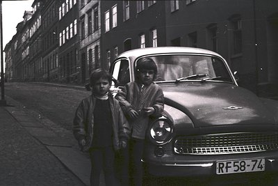 Mid-century classic car (likely a Citroën DS) parked beside two boys in urban setting. Uniform brick apartment blocks and vin...