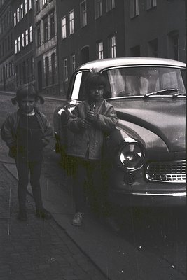 Black-and-white street scene featuring two men in 1950s-era attire posing beside a vintage car. The man on the left wears a f...