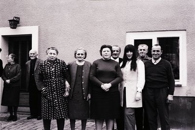 Black-and-white group portrait of 8 adults posing outdoors in front of a residential building, likely from the 1960s–1970s. F...