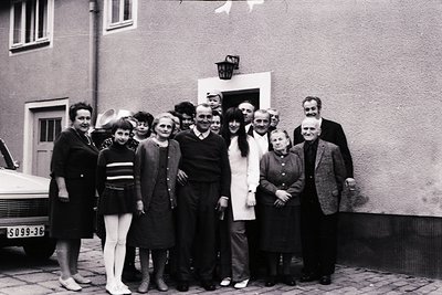 Nine adults pose outside a residential building in mid-20th century attire, likely 1950s–1960s. Women wear knee-length dresse...