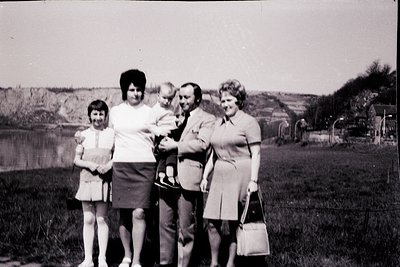 Family portrait from the 1960s–70s, likely Eastern Europe. Five individuals pose outdoors near a body of water and rural land...