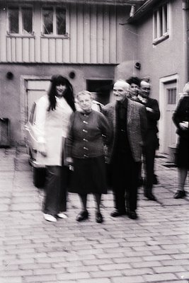 Blurred group photo in a European courtyard, likely 1970s–1980s. Three central figures in mid-motion, surrounded by older adu...