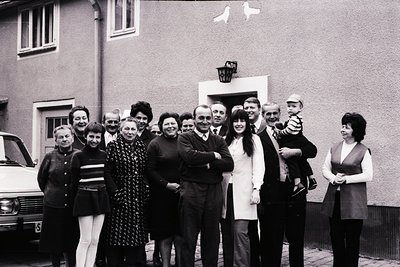 Family portrait in front of a residential building, likely 1960s–1970s. Nine adults and a baby pose outdoors, dressed in mid-...