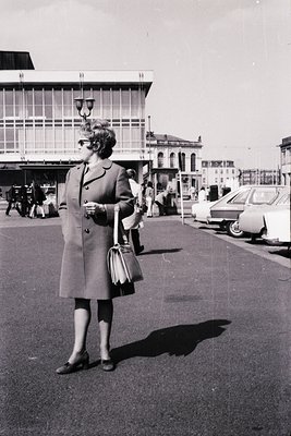 Mid-century woman in a knee-length coat and gloves stands near a modernist bus terminal, holding a small handbag. Classic 196...