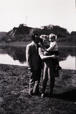 Family portrait by a serene lake, framed by grassy shore and distant rocky hill. Man in suit, woman in high-waisted skirt, ch...