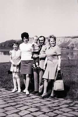 Family portrait on paved pathway beside a lake, 1960s. Four adults and a baby in matching mid-century attire—pleated skirts, ...