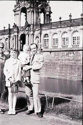 Baroque-style palace façade with ornate columns and arched windows in background. Family portrait: man in light suit, woman i...