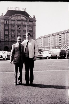 Two men in formal attire pose in front of a Soviet-era building labeled "CENTRUM" in Moscow, USSR, circa 1960s–1970s. The arc...