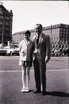 Two individuals pose in formal attire on a city street, likely 1960s. The woman wears a knee-length jacket, white tights, and...