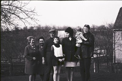 Six individuals pose outdoors in a 1960s-70s suburban setting. Men wear suits/coats, women in knee-length dresses and cardiga...