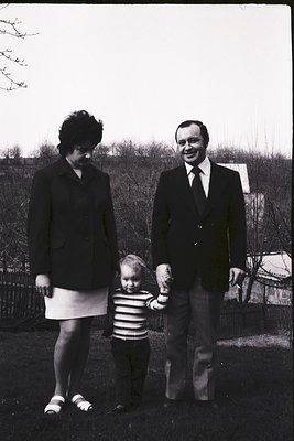 Family portrait in 1960s-70s suburban setting. Woman in fitted blazer and skirt, man in suit with white shirt, child in strip...