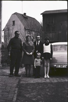 Family portrait in 1950s-60s European residential setting. Five individuals pose on a cobblestone street beside a vintage Cit...