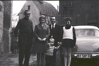 Family portrait in 1960s European countryside. Four adults and a toddler pose outdoors beside a Citroën DS (RF58-7L plate). M...