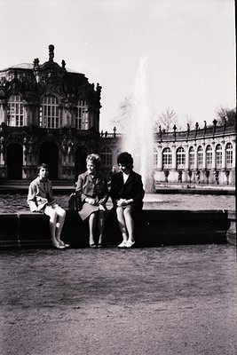 Three individuals pose near a Baroque-style fountain at the **Sanssouci Palace**, Potsdam, Germany. The man sits left in a sh...