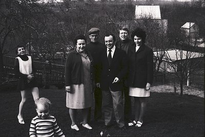 Group portrait in mid-20th century outdoor setting, likely 1950s–1960s. Seven individuals pose in a park-like area with leafl...
