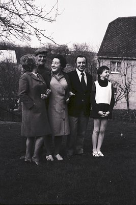 Four individuals pose outdoors in 1960s European suburban style—men in suits, women in knee-length coats and dresses. Leaves ...