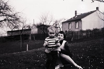 Two children in a mid-20th-century backyard, likely 1950s–1960s. The girl, holding a younger sibling, wears a striped sweater...