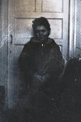Vintage black-and-white portrait of a person standing in a dimly lit hallway, partially obscured by a frosted glass door. The...