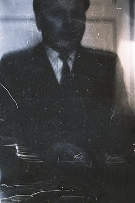 Silhouetted figure in a dark suit and tie, reflected on a textured, possibly metallic surface. The blurred background suggest...