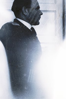 Silhouetted figure in a dark overcoat and white shirt, captured through rain-streaked glass. Mid-20th century urban streetwea...