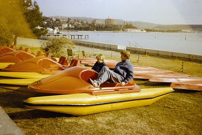 Two children sit on a vintage pedal boat by a lakefront, surrounded by matching orange/yellow pedal boats. Mid-20th century r...