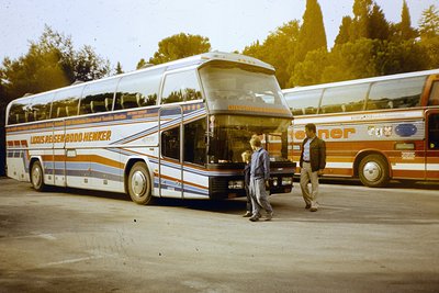 Two vintage tour buses parked side-by-side in a lot, 1970s. The bus on left reads "Luxus Reisen Budg Henker" with orange/whit...