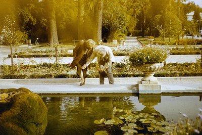 Two individuals tending to a pondside planter in a formal garden, likely mid-20th century. The man in a light-colored suit an...